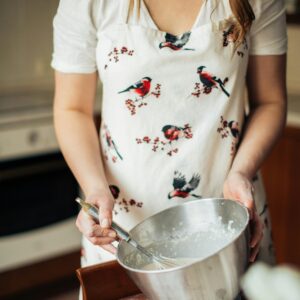 A woman in a kitchen mixing dough in a bowl while wearing a bird-patterned apron.